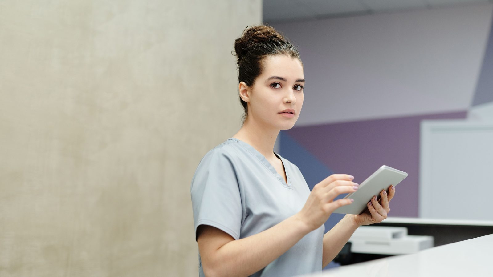 Profissional de saude em pe no balcao de uma recepcao de clinica, consultando um tablet.