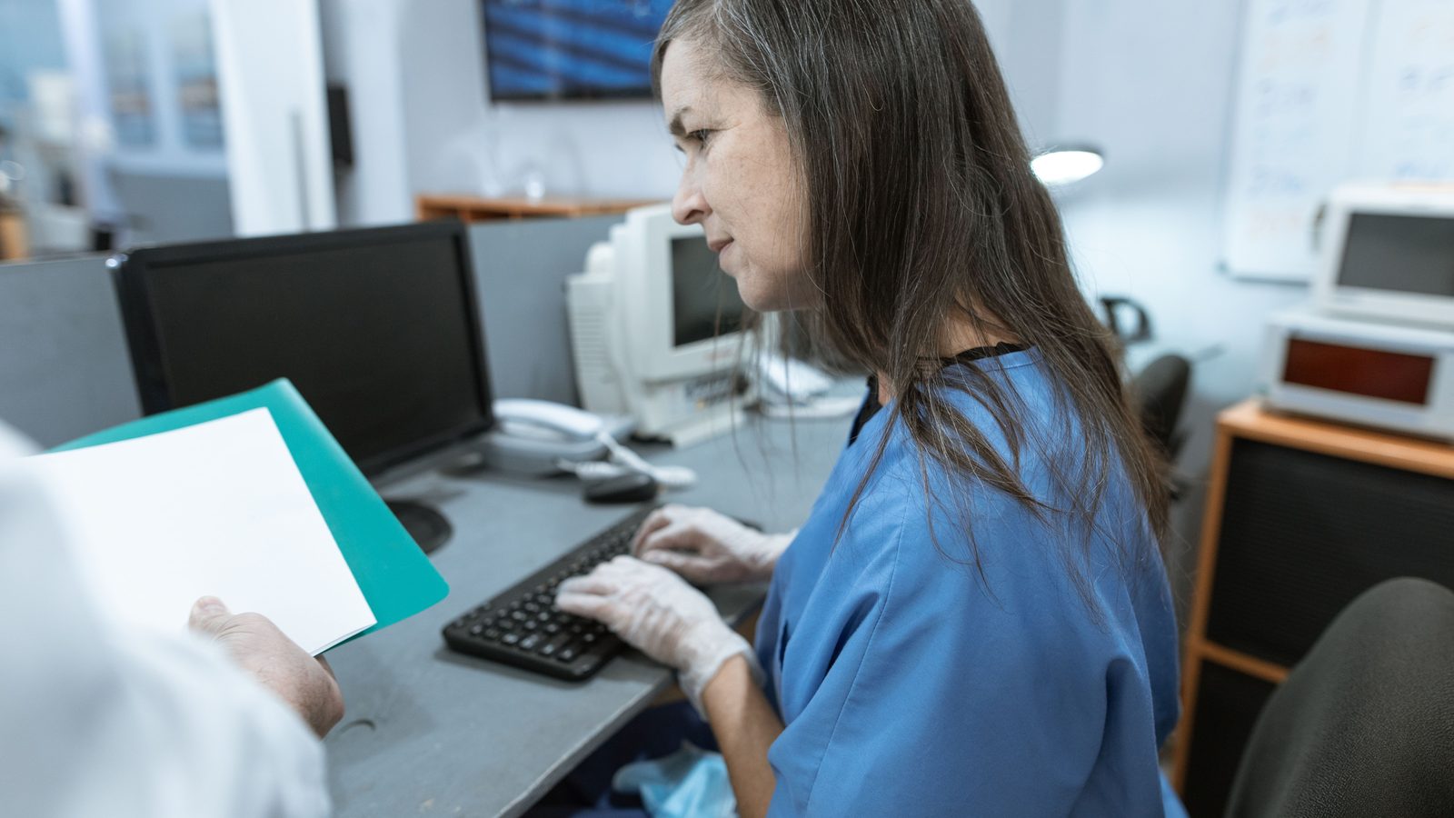 Profissional de saúde em um computador de recepção, com uma pasta de documentos sobre a mesa.