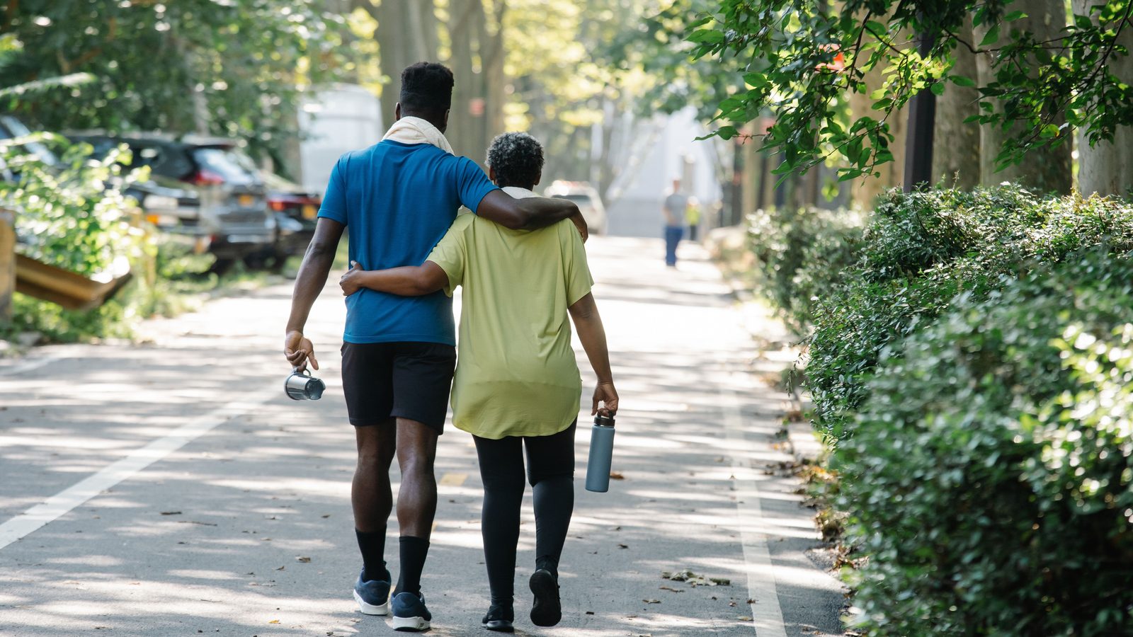Filho adulto caminhando de braço dado com a mãe idosa em uma via arborizada.