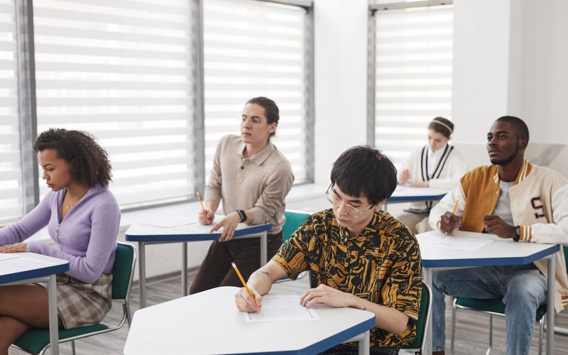 Estudantes fazendo prova em carteiras de uma sala de aula iluminada.