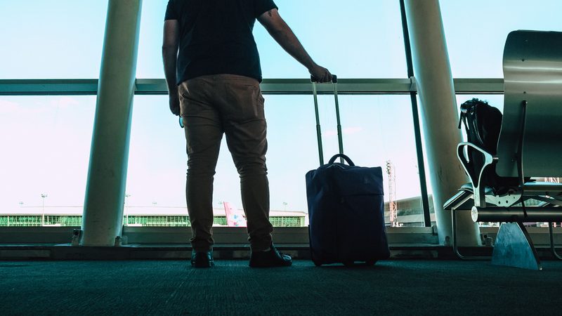 Homem de costas com mala de viagem diante da vidraça de um aeroporto.