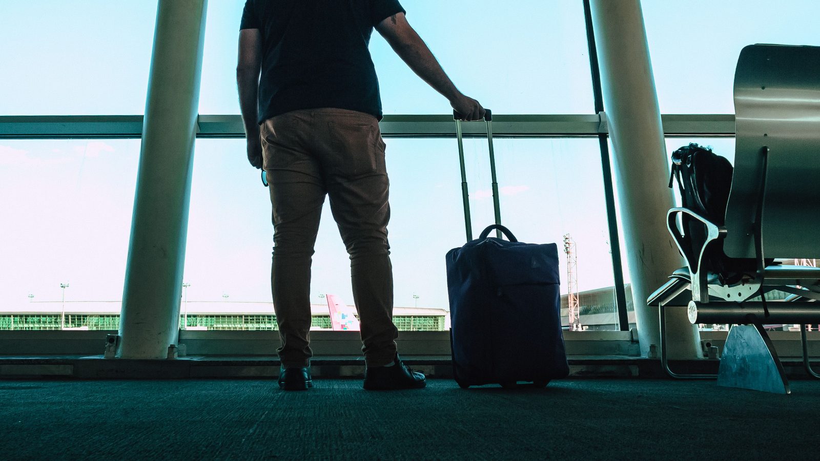 Homem de costas com mala de viagem diante da vidraça de um aeroporto.