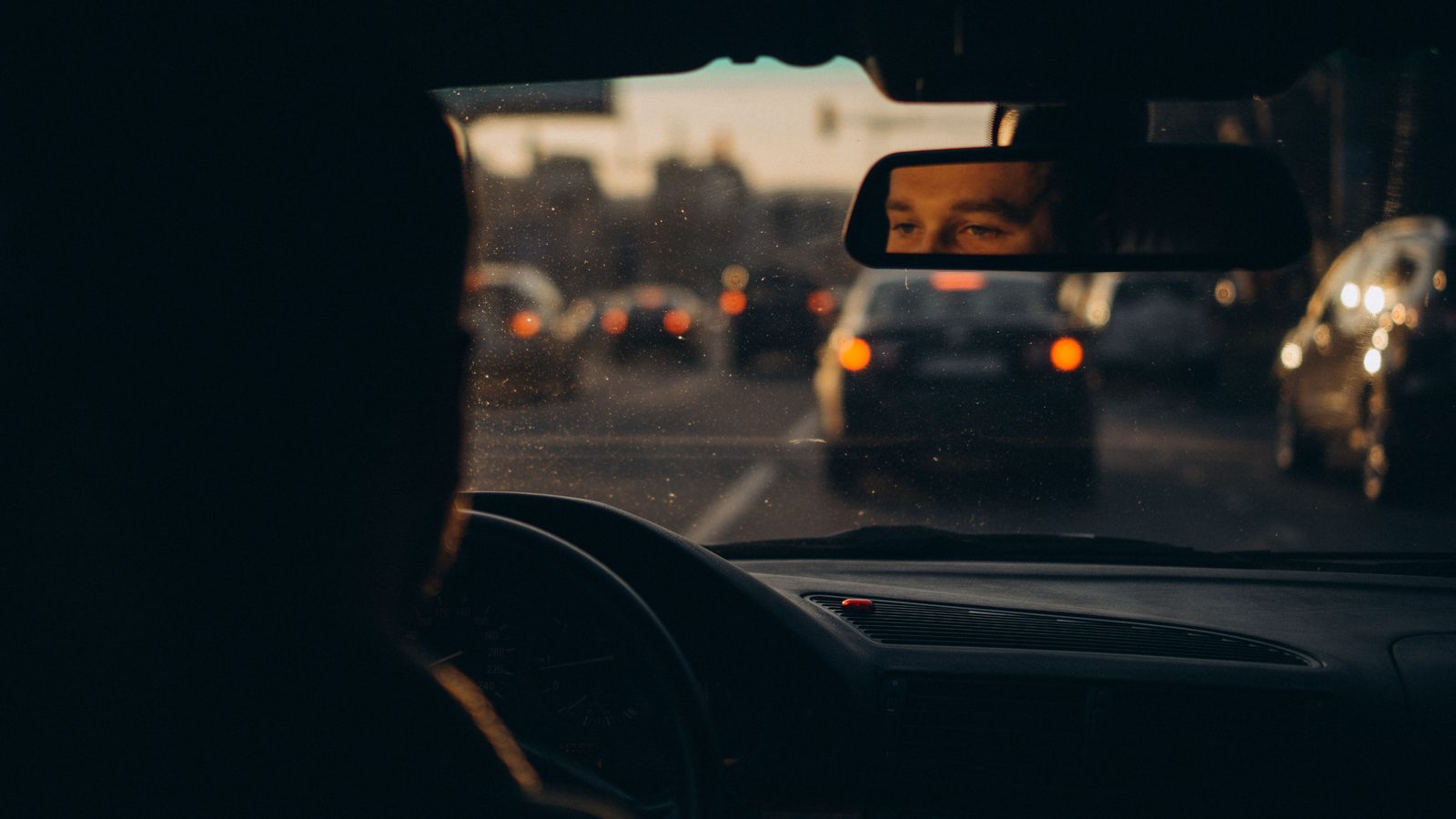 Motorista refletido no espelho retrovisor enquanto dirige em trânsito no fim do dia.
