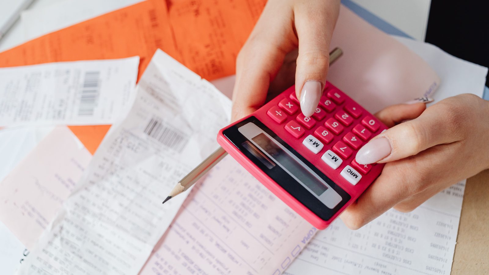 Mãos usando uma calculadora sobre uma mesa coberta de comprovantes e papéis de contas.