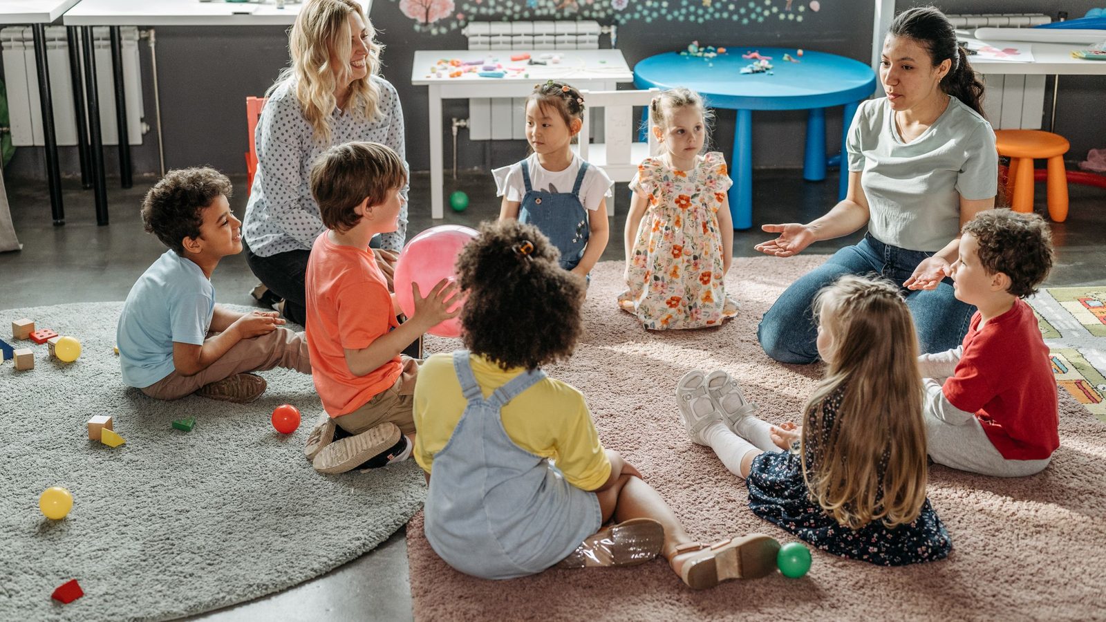 Crianças pequenas sentadas em roda no chão de uma sala de creche, com duas educadoras conversando com elas.