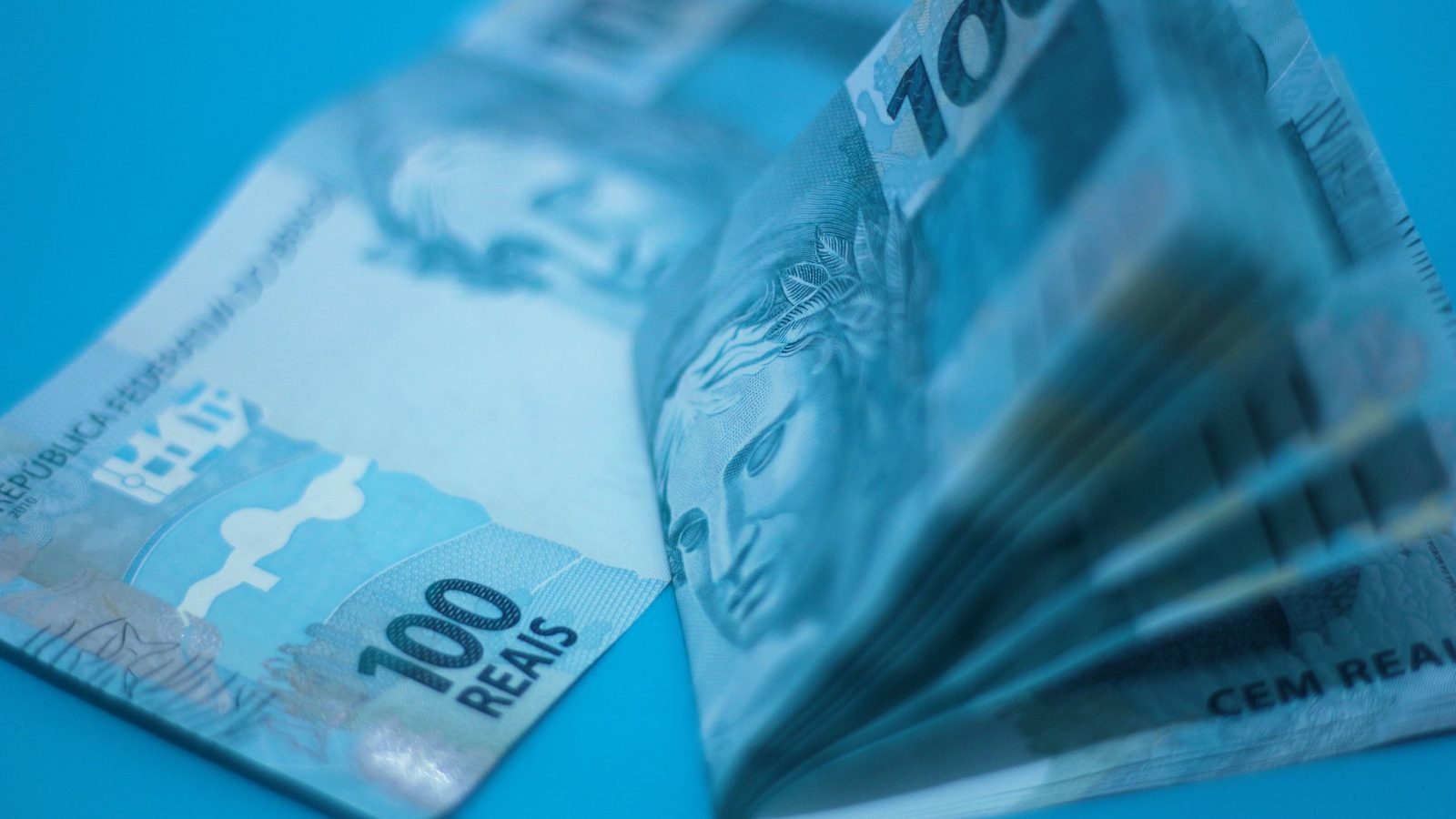 Notas de cem reais empilhadas sobre uma superfície azul.