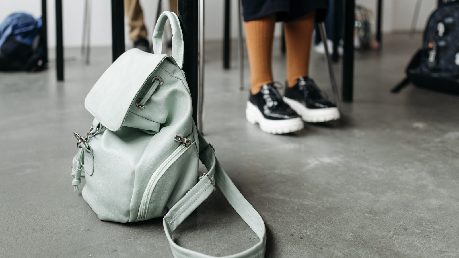 Mochila verde apoiada no chão de uma sala de aula, com carteiras ao fundo.