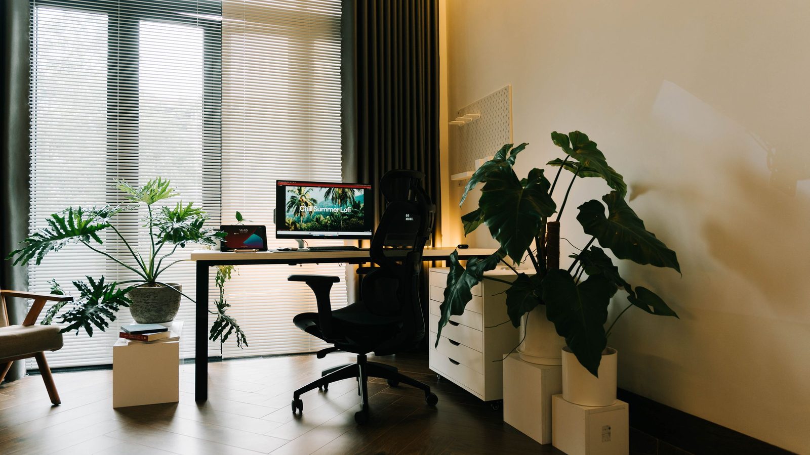 Escritório montado em casa, com mesa junto à janela ampla e plantas ao lado.