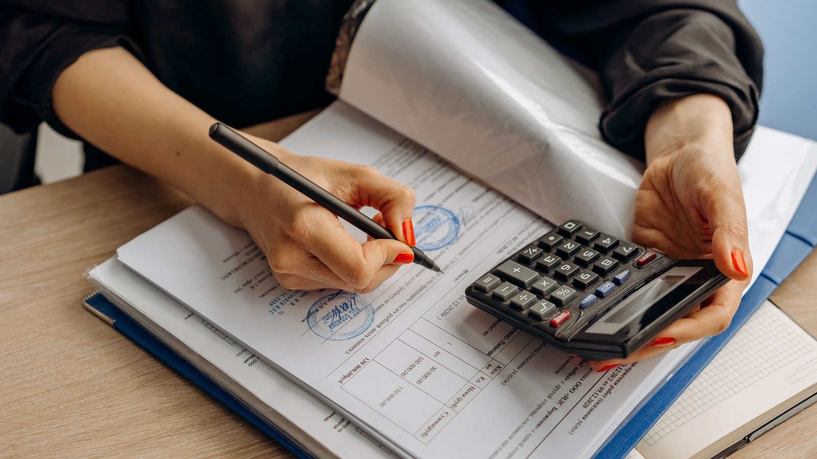 Pessoa usando calculadora e assinando papéis sobre uma mesa com documentos e pastas.