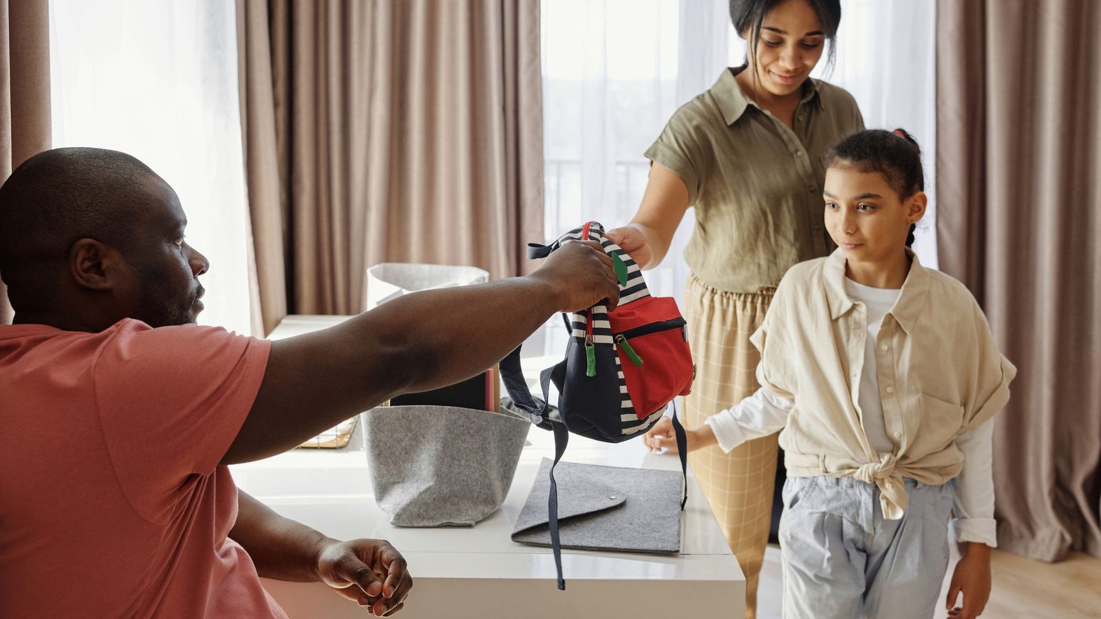Pai, mãe e filha preparando a mochila escolar sobre uma mesa em casa.