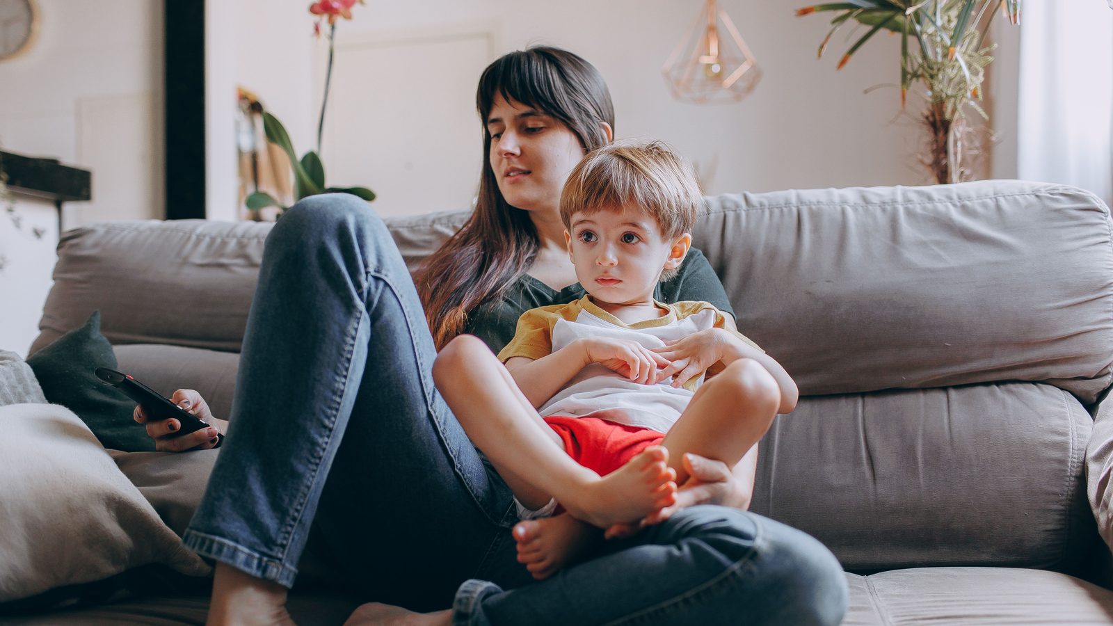 Mãe e criança pequena sentadas juntas no sofá de uma sala clara, em casa.