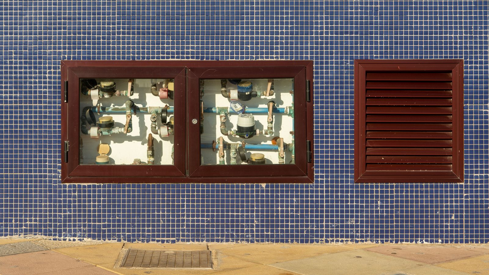 Caixa de hidrômetros com registros e canos aparentes na fachada de pastilha azul de um prédio.